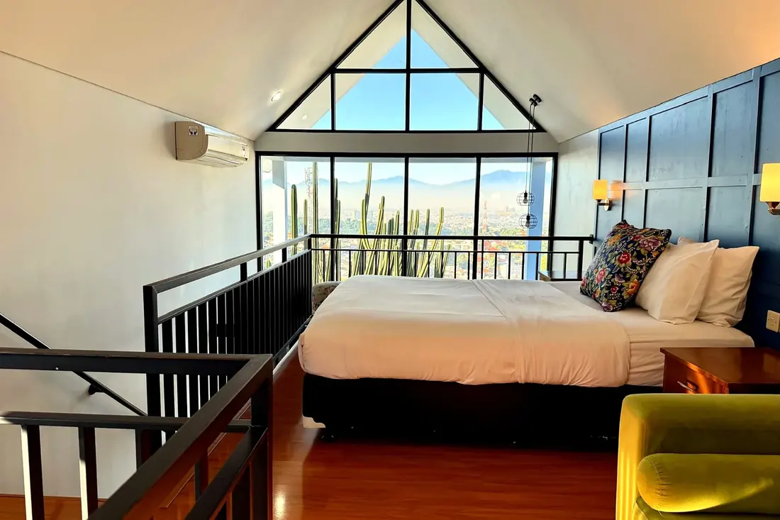 Mezzanine bedroom with blue paneled feature wall, queen bed, lime green sofa and triangular picture windows overlooking Bandung city and mountains